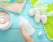 A hand holds a baby bottle filled with milk. Nearby are a can of baby formula with a scoop, a pacifier, a green bib with a crochet horse toy, and a soft towel. The background is a turquoise wooden surface.