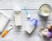 Baby bottles filled with milk and covered with caps rest on a light wooden surface, accompanied by a small blue spoon, a white towel, a can of powdered formula, a spoon with an orange handle, and a plush toy dog.