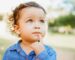 A toddler with curly hair and earrings is looking up thoughtfully, a finger on their chin, perhaps pondering the choice between breast milk or the right infant formula. They are wearing a blue denim shirt against a softly blurred backdrop of greenery and a building.